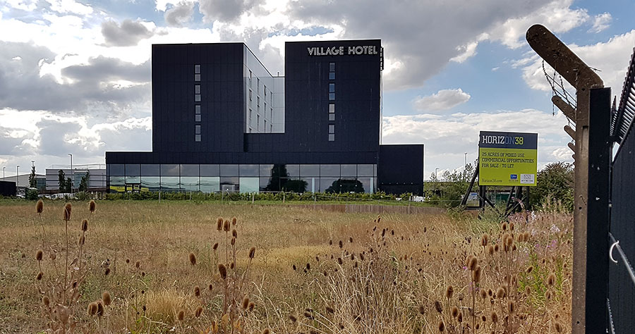 Photo of overgrown vacant land with a hotel in the background.